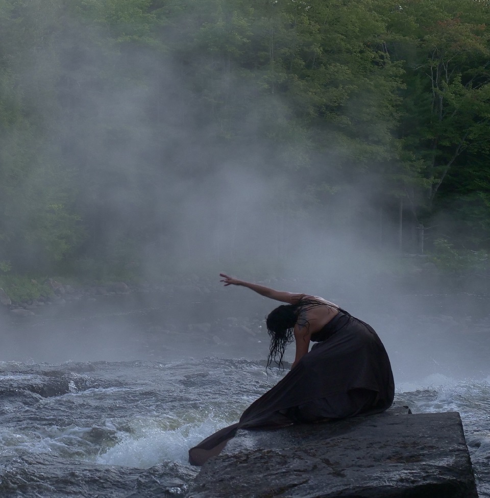 A dance artist leans over on a rock in front of rushing water. Their hair is loose and wet and they have on a long black dress.