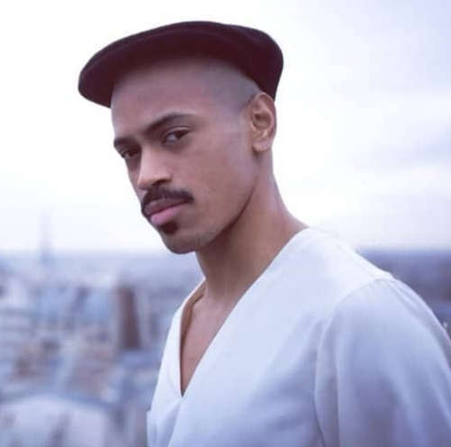 A young Black man wearing a beret and white T-shirt looking directly at us, with a city vista in the distance behind him