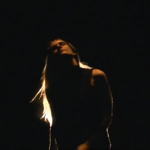 A dance artist sways in a black room. Their head is pointing up to the sky and their long blond hair is lit up by a light outside of the image.