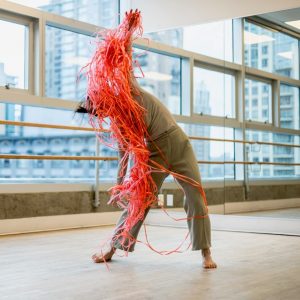 A dance artist wearing tan pants and shirt leans to the side in a bright dance studio. They are holding a large bundle of red string which is obscuring their face and is spread out down the length of their body.