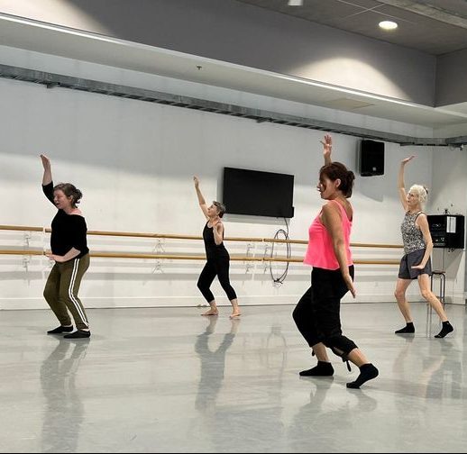 Four dancers in a bright studio. They are all wearing rehearsal clothing and are facing towards the mirror, slightly away from the camera. They have their right arms up in the air and their knees slightly bent. Their body's are slightly arched in a exuberant fashion.