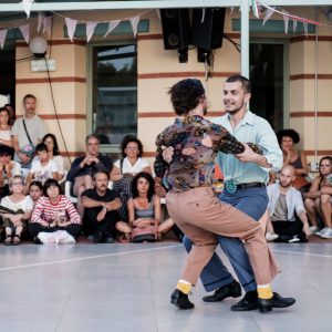 Two men mid dance. They are holding on to each others arms tightly and their knees are interlocked as if they are about to spin around. They are smiling and oath wearing trousers and button up shirts They are being watched by a crowd sitting on the floor and chairs in a circle.