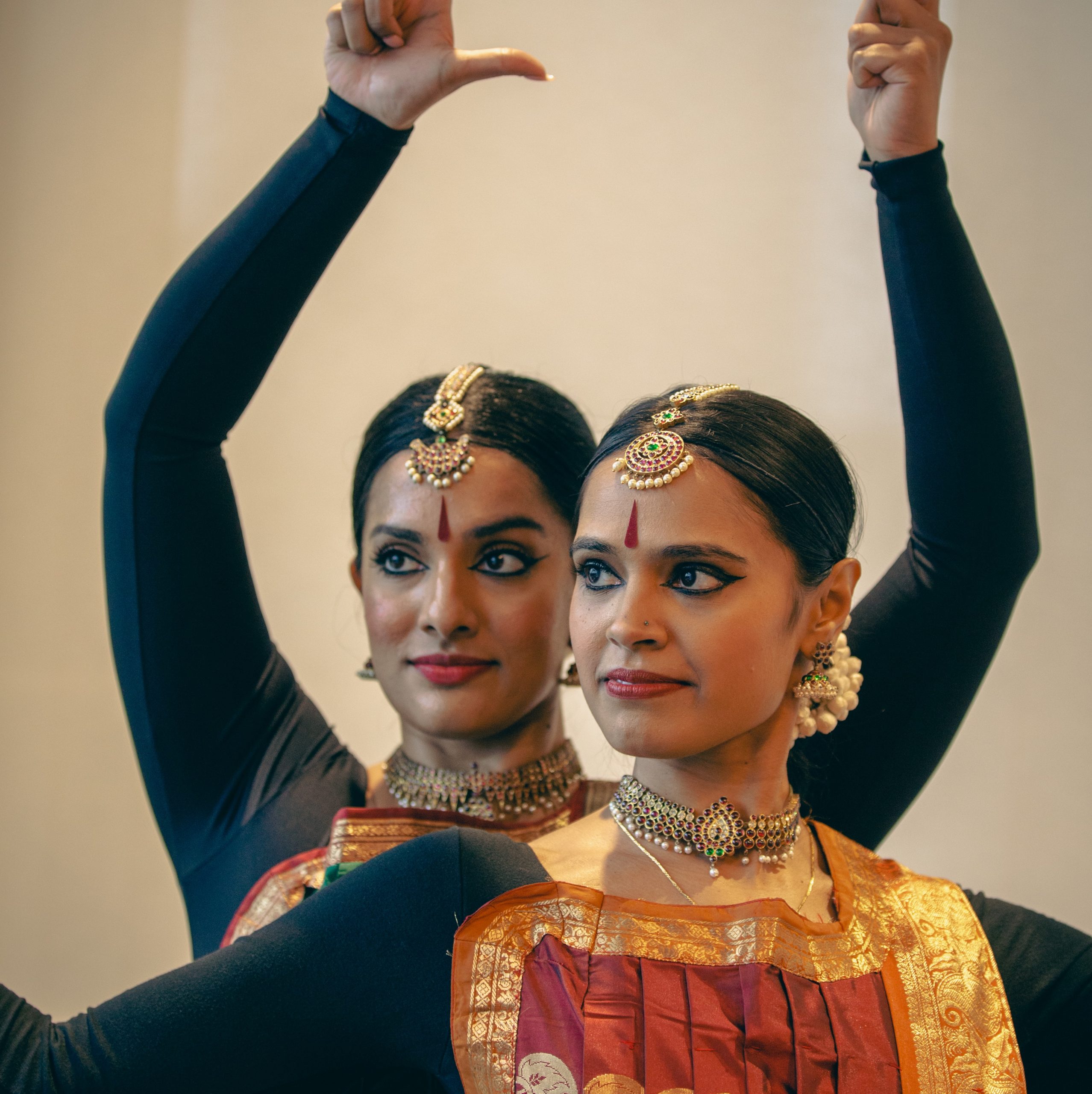 Two women in traditional bharatanatyam clothing, hair, makeup and jewelry pose for the camera. The woman in the back has her arms raised with thumbs pointing out.