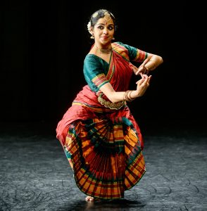Shilpa Shankar poses in a black theatre wearing a traditional bharatanatyam outfit of red, teal and orange. Her arms are outstretched with her hands linked and she is looking just to the side of the camera.