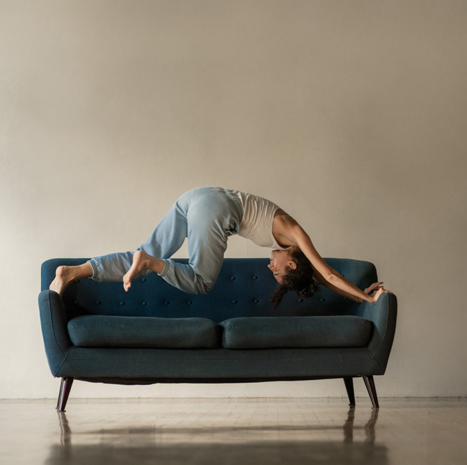 A dance artist leans over a blue couch with their feet on one arm and their hands on the other. They are wearing a white tank top and blue sweatpants. The couch is also blue but a darker shade. They are in a beige room with a shiny floor and a plain beige wall behind them.