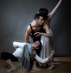 Three people pose together. One is crouched in a low squat and is leaning over slightly to the left. The middle person is wearing a black tank and is bent slightly at the waist. The third person is standing with their back to the camera, leaving on the middle person. They are wearing a long white dress and have their left arm in the air.