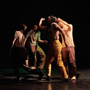 Four dance artists move in a circle in a black theatre. They are wearing casual neutral clothes and are all moving their arms and torsos.