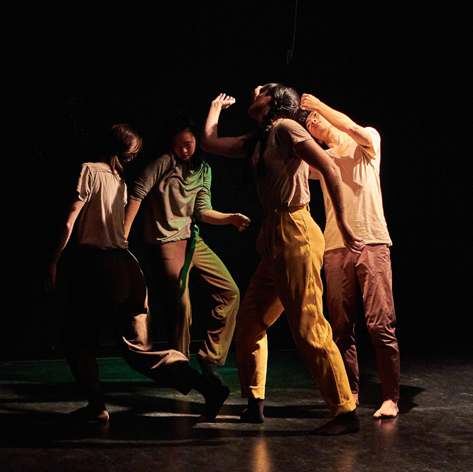 Four dance artists move in a circle in a black theatre. They are wearing casual neutral clothes and are all moving their arms and torsos.