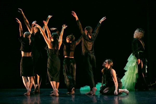 Dance artists on a dark stage mid performance. Five are turned away from the camera with their arms up. One sits on the floor looking up at them and another walks away. They are all wearing black and the one walking away has a long fluffy green skirt trailing behind them.