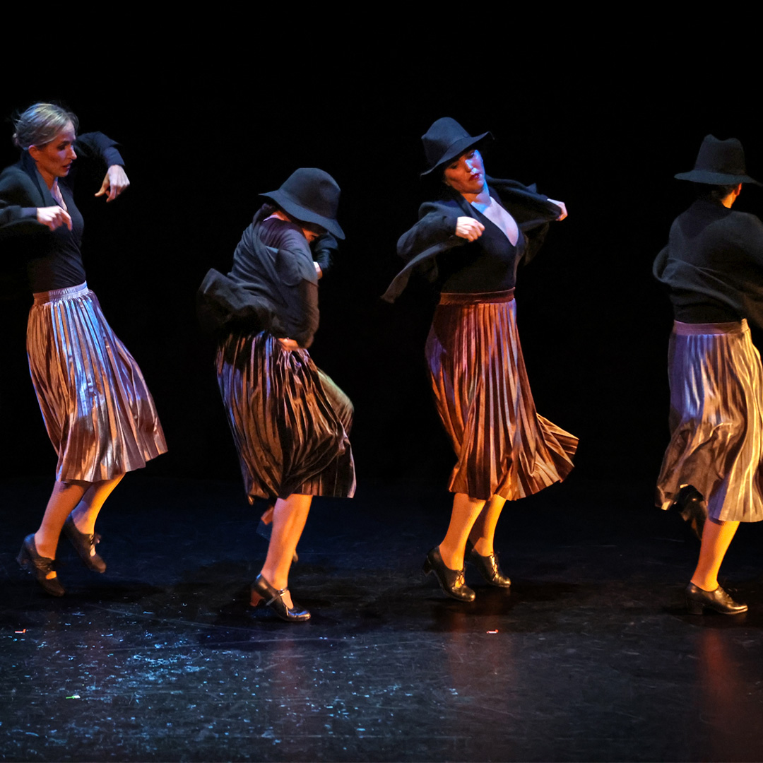 Four dance artists mid performance on a dark stage. they are all wearing pink skirts, black tops and jackets and three have black floppy hats. They are standing in a line across the stage.