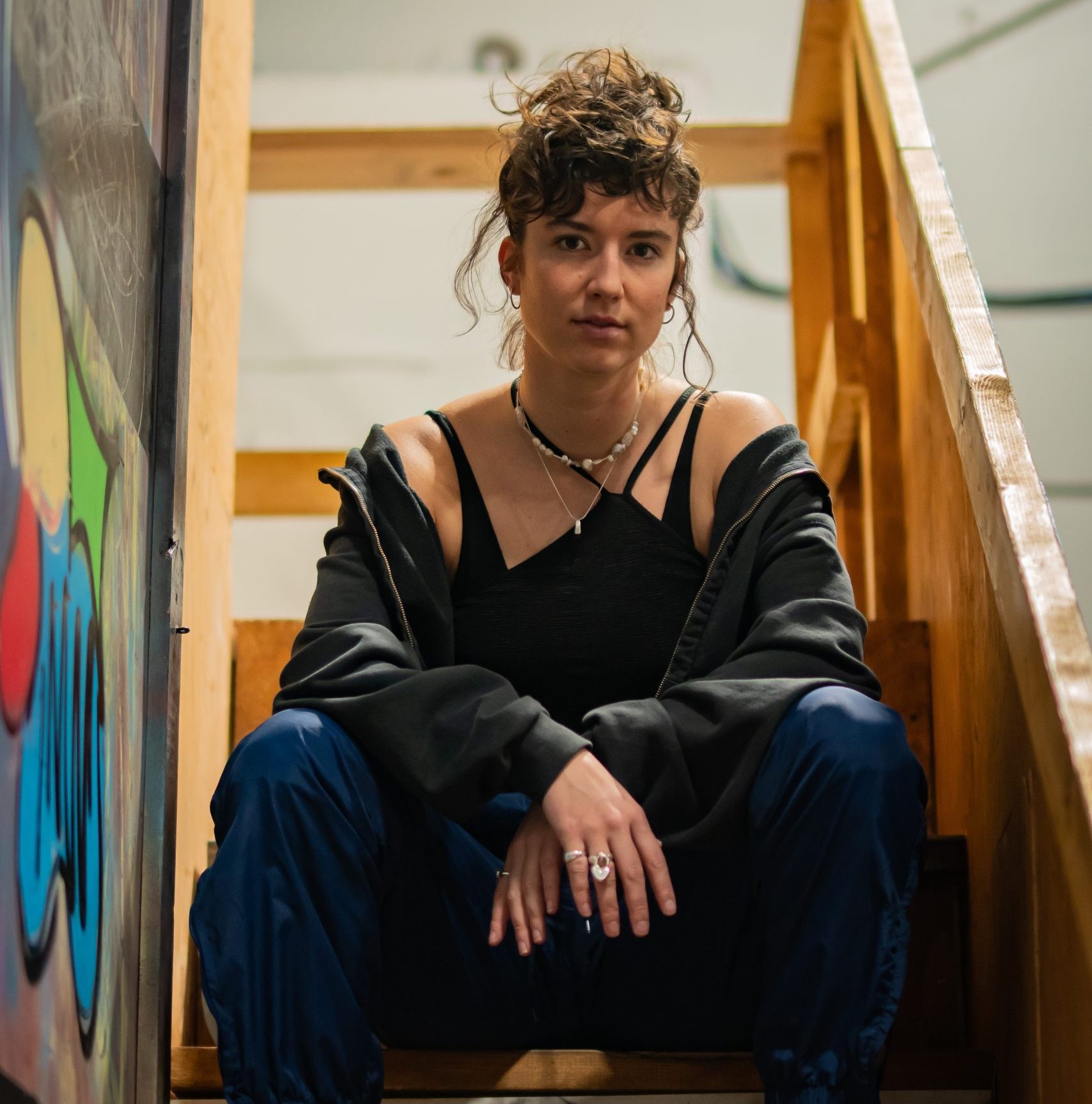 Head shot of Anya Saugstad: a woman sits on wooden steps. Her curly dark hair is up and she is wearing a black sweater that is falling off her shoulders showing her necklaces and black tank top.