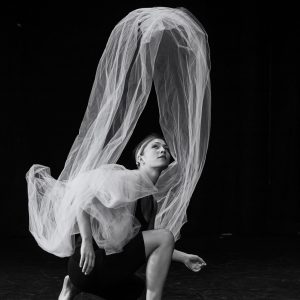 A dance artist kneels on a black theatre floor, looking up and behind them at a large piece of tulle that is twisting in the air. This image is in black and white.