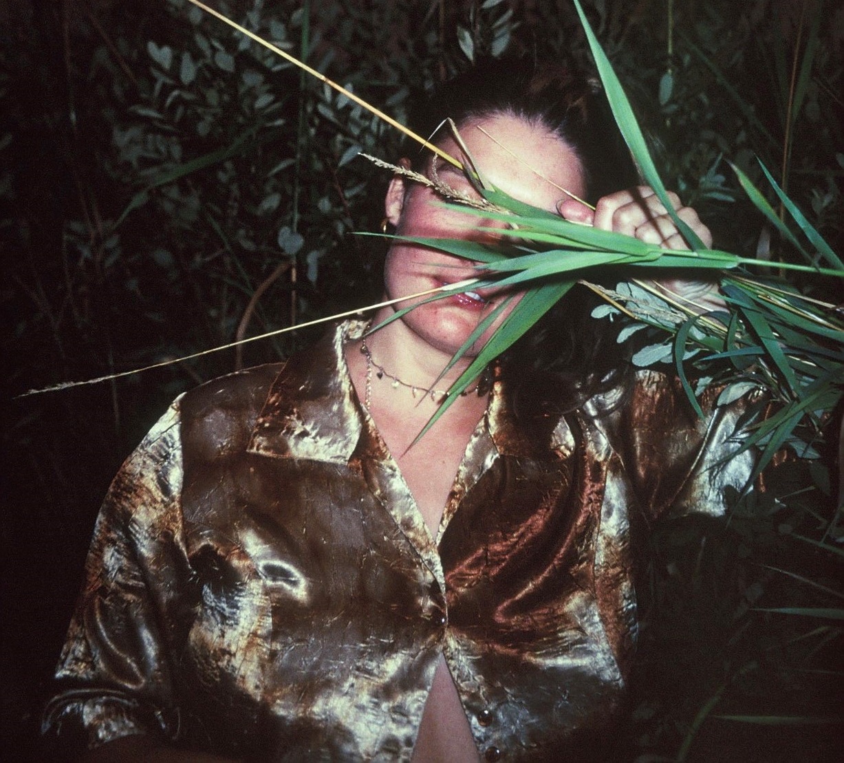 A dance artist in a shiny brown shirt holds plant branches and leaves in front of their face. There are more plants behind them.