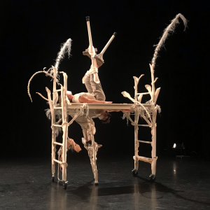A white structure stands in the middle of a black stage. It has ladders as it's legs and a plank across the top. There are long white sticks and feathers sticking out the top. Two dance artists interact with the structure - one hanging from the plank with one leg on the ground and the other lying on the top of the plank with legs in the air. Both dance artists are wearing white and have stilts on.