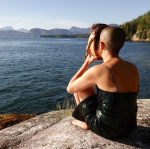 A dance artist on a rock wearing a sparkly black dress sits facing the ocean, away from the camera, holding a mask up to their face