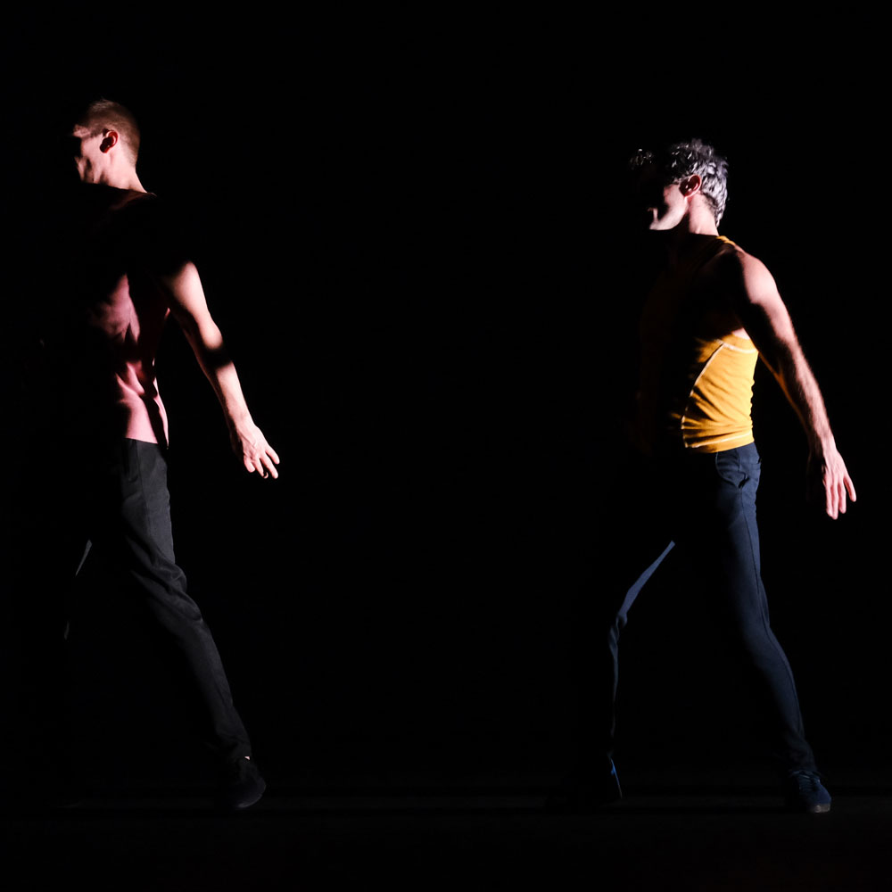 Two dance artists only slightly illuminated pose on a dark stage. They are standing with one leg slightly bent and arms extended downwards.