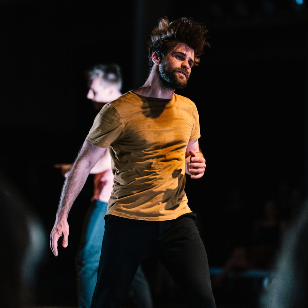 A dance artist mid jump. Their hair is flying and their yellow shirt has sweat marks on it. There are other dance artists behind them.