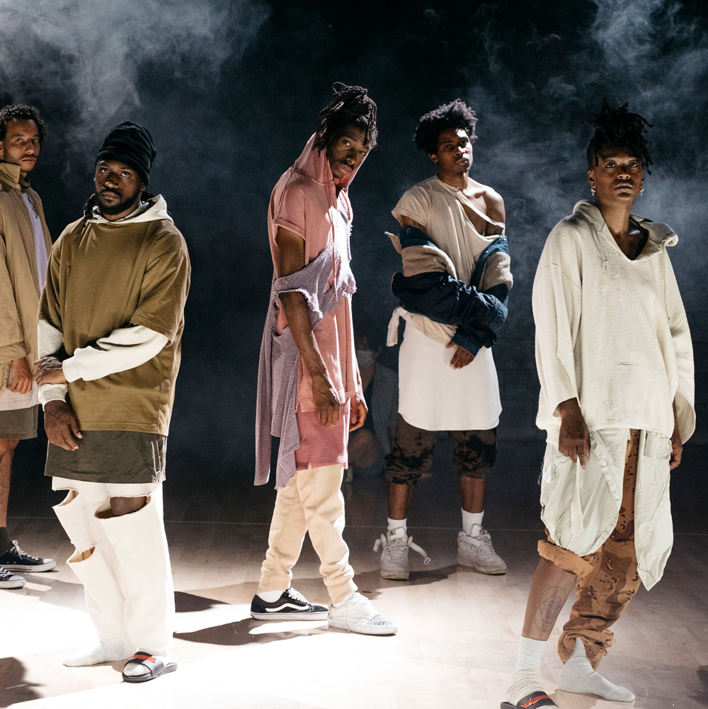 Five dance artists stand looking at the camera. They are wearing baggy clothes, some of which that are torn. There is smoke behind them.