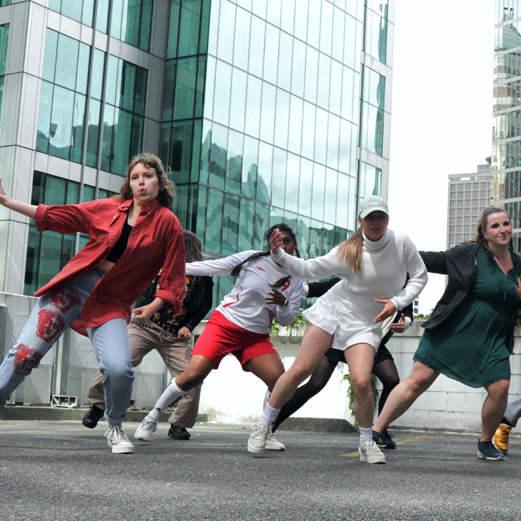 A group of dance artists mid performance on a rooftop patio in a city. They are all wearing street clothes and are lunging to the side with their arms reaching out.