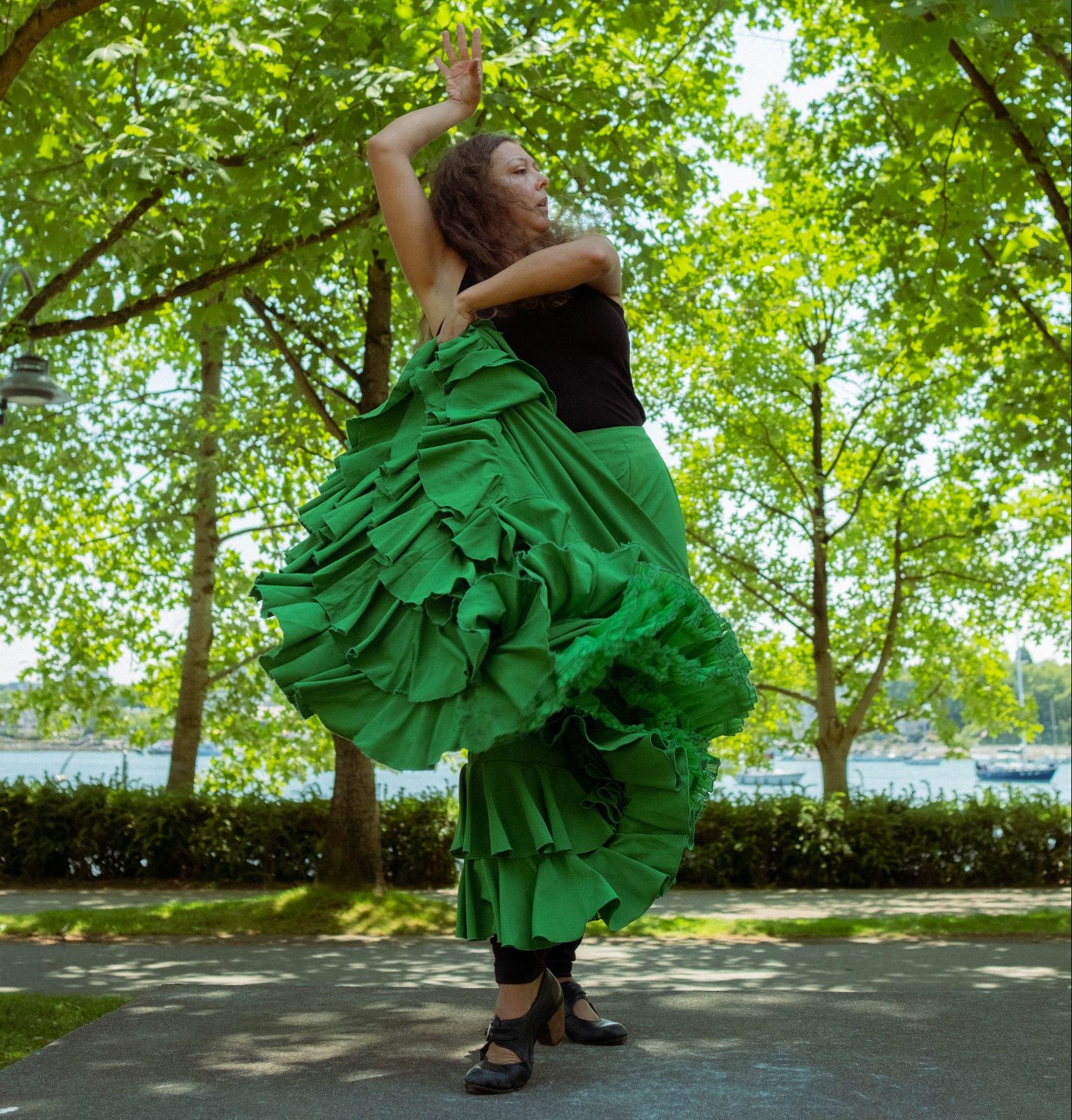 A flamenco dancer poses in a park. She is wearing a green traditional skirt that she is holding up with her left arm. Her right arm is bent above her head and she is looking out to the left.