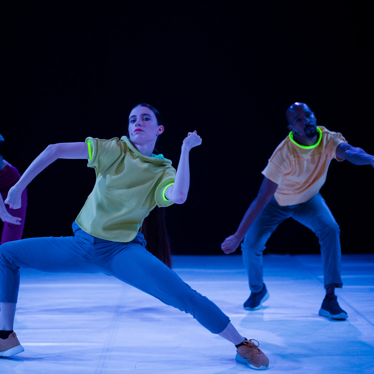 Two dance artists wearing bright clothes pose mid performance. They have glow sticks around their necks and arms and are wearing bright running shoes on the white dance floor.
