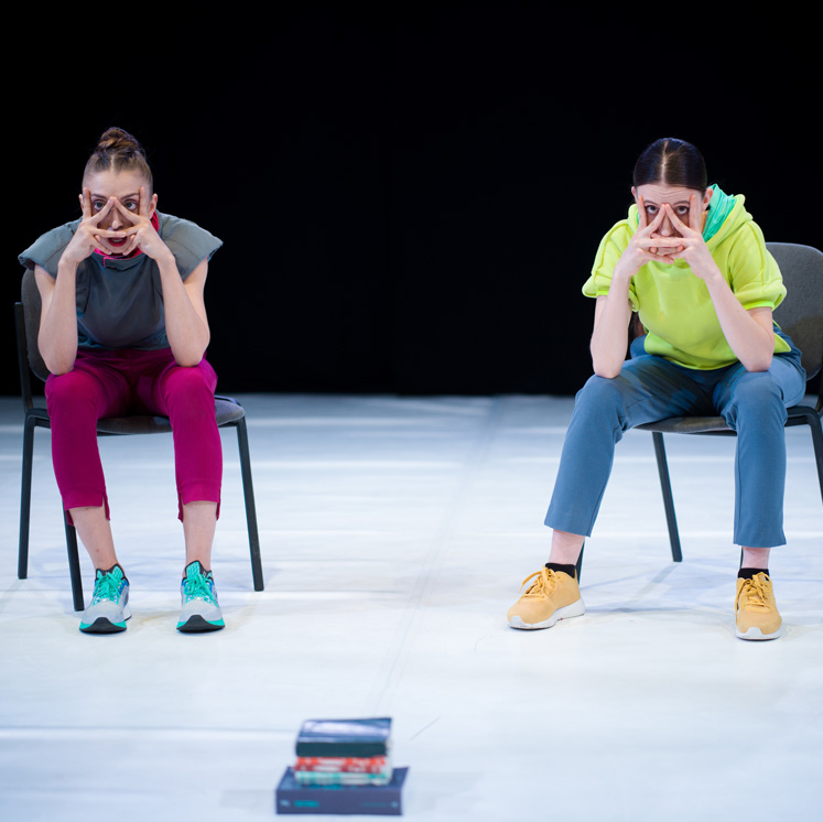 Two dance artists sit on office chairs mid performance. They are wearing bright clothing and sneakers and have their fingers up around their face so their eyes and noses are poking out.