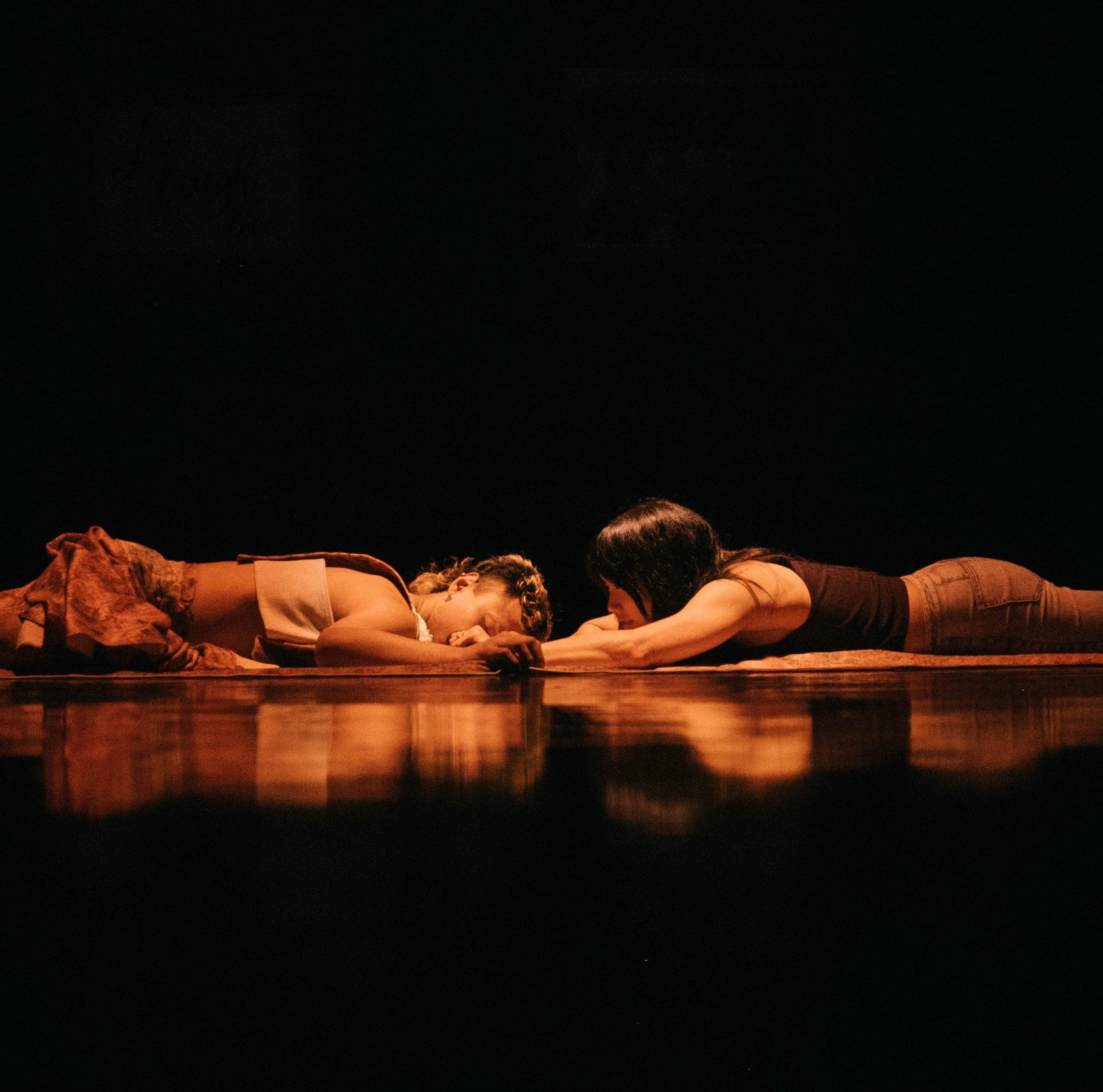 Two dance artists lie on their stomachs facing each other on a dark dance floor. There is a warm light shining on them and one has their had down with eyes closed as if sleeping and the other is looking and reaching towards the first.