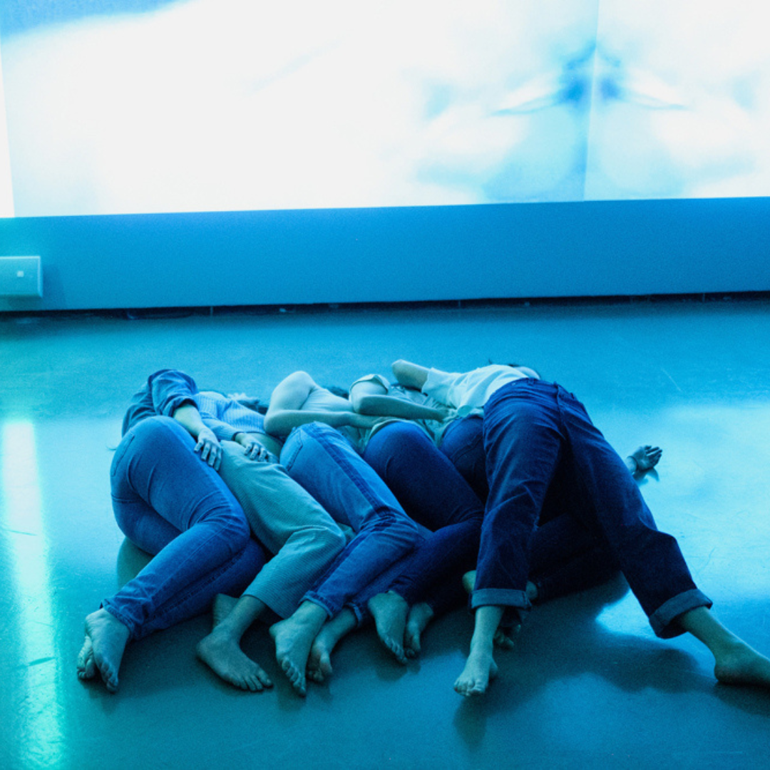 Six dance artists lie on a floor. Five of them are spooned together and one is rolling over top of the others. They are all wearing blue jeans. The lighting in the room has a blue wash.