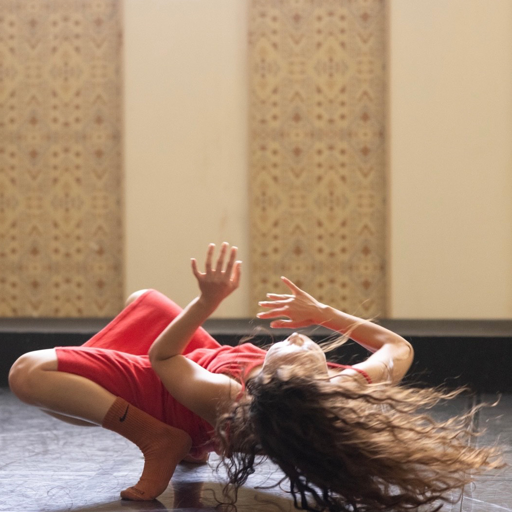 A dance artist balances on their toes as they hold their body just above the floor. They have long curly hair and are wearing a red dress.