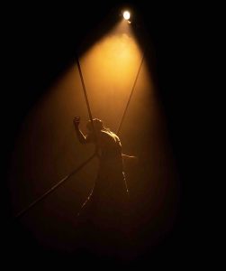 A dance artist leans against suspended ropes. the artist is arched back with one hand up, fingers clenched. There is a solo spotlight on and smoke around giving the light an orange glow amidst the darkness.