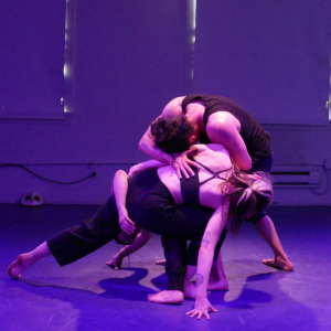 Two dance artists lean down, one over the other. they are wearing rehearsal clothing and facing away from the camera.