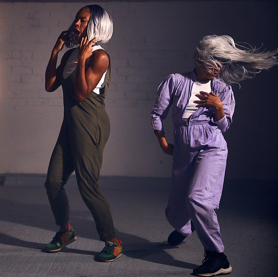 Two dance artists mid dance in a dark room. they are wearing jumpsuits - one olive green, one purple and have silver/grey wigs on.
