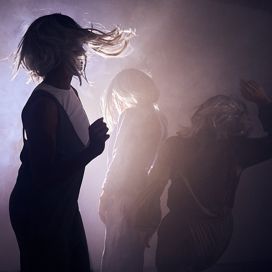 Three dance artists wearing silver wigs dance amidst a bright light and smoke.