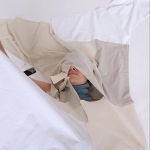 A person lies on white fabric with tan fabric covering their face and torso. Through a hole near their neck you can see a blue turtleneck. There is also more white fabric coming down onto the artist and covering the rest of their body,