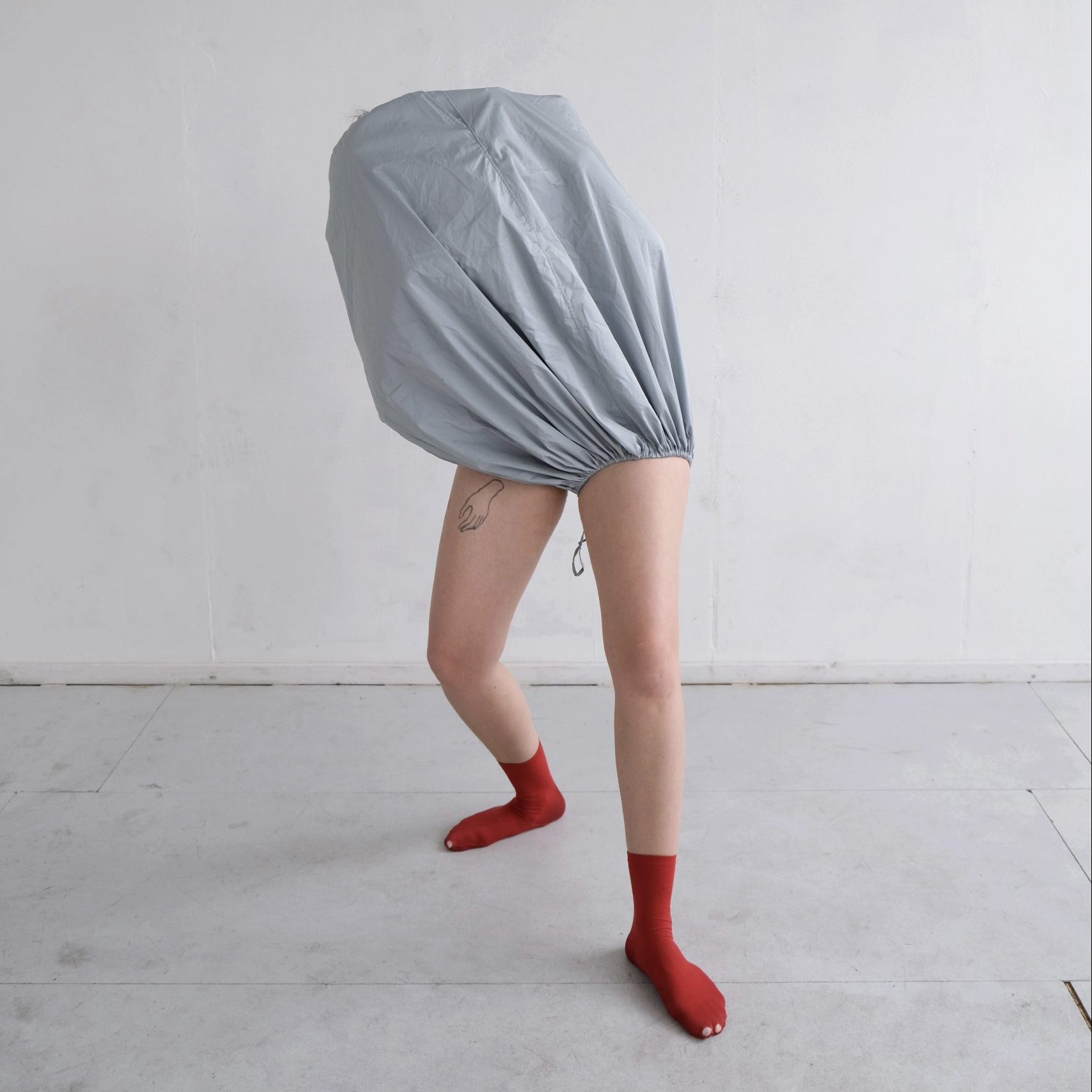 A dance artist poses in a grey studio. They have grey fabric covering their entire head and torso and are wearing bright red socks.