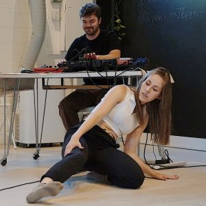 A dance artist sits, leaning on their left arm while extending their right leg. Behind them a technician works on a sound board sitting on a table.