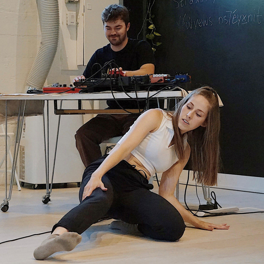 A dance artist sits, leaning on their left arm while extending their right leg. Behind them a technician works on a sound board sitting on a table.