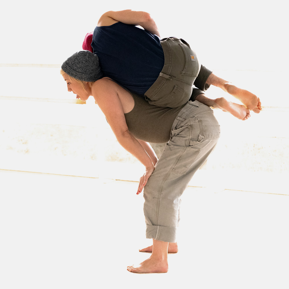 Two dance artists in a bright space. One is bent over with their hands on their knees and the other is balancing on the firsts back curled in a ball. They are both wearing tank tops, toques and khakis.