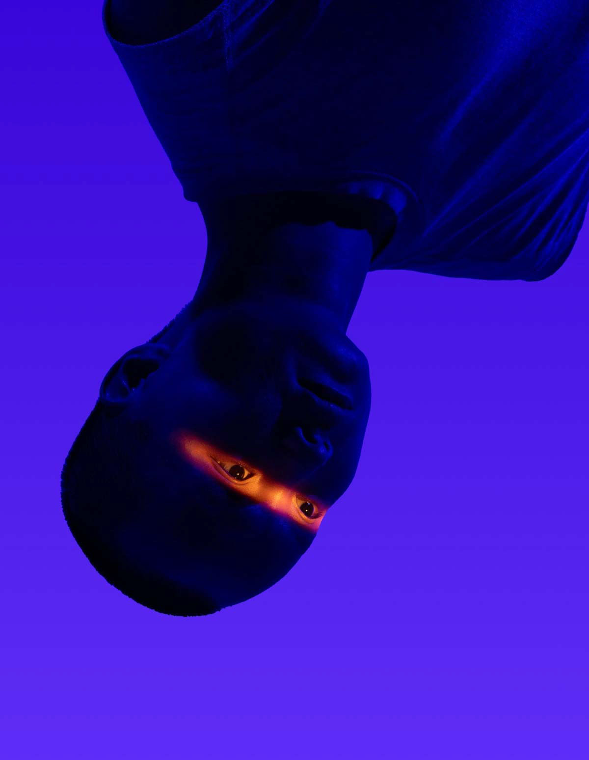 A dance artist is lit in all blue except for a bright light shining across their eyes. The artist appears to be hanging upside down.