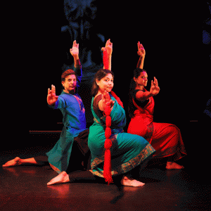 Three dance artists pose on a dark stage. They are all one one knee with their arms outstretched, above and in front of them and they are looking at the camera.