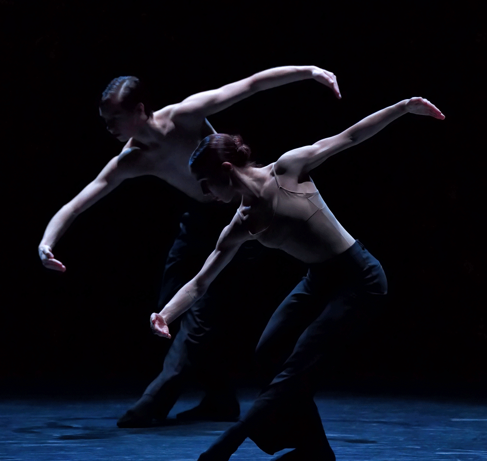 Two dance artists post on a dark stage. They are leaning forward with one leg and their arms extended in ballet fashion. They are wearing black pants.