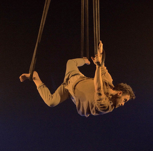 An aerial dancer hangs from multiple ropes in the dark. He is wearing tan pants and a long sleeve shirt and his body is twisted in multiple directions.