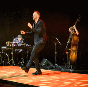 A tap dance artist mid performance - he is kicking one leg up. Behind him are two musicians one playing a piano and percussion, the other with an upright bass.