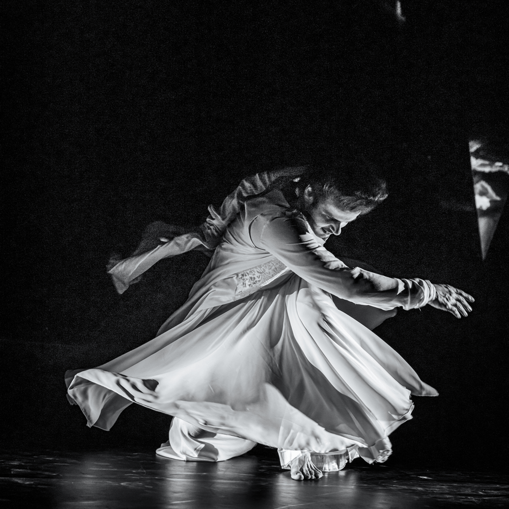 A dance artist crouches down on a dark stage. He is wearing flowing clothing that flows around him as he moves. His arms are extended as if about to twirl.
