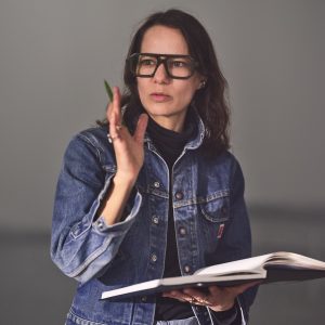 Headshot of Justine Chambers, looking just past the camera. She is wearing thick glasses and is holding a book open in one hand and a pen in other.
