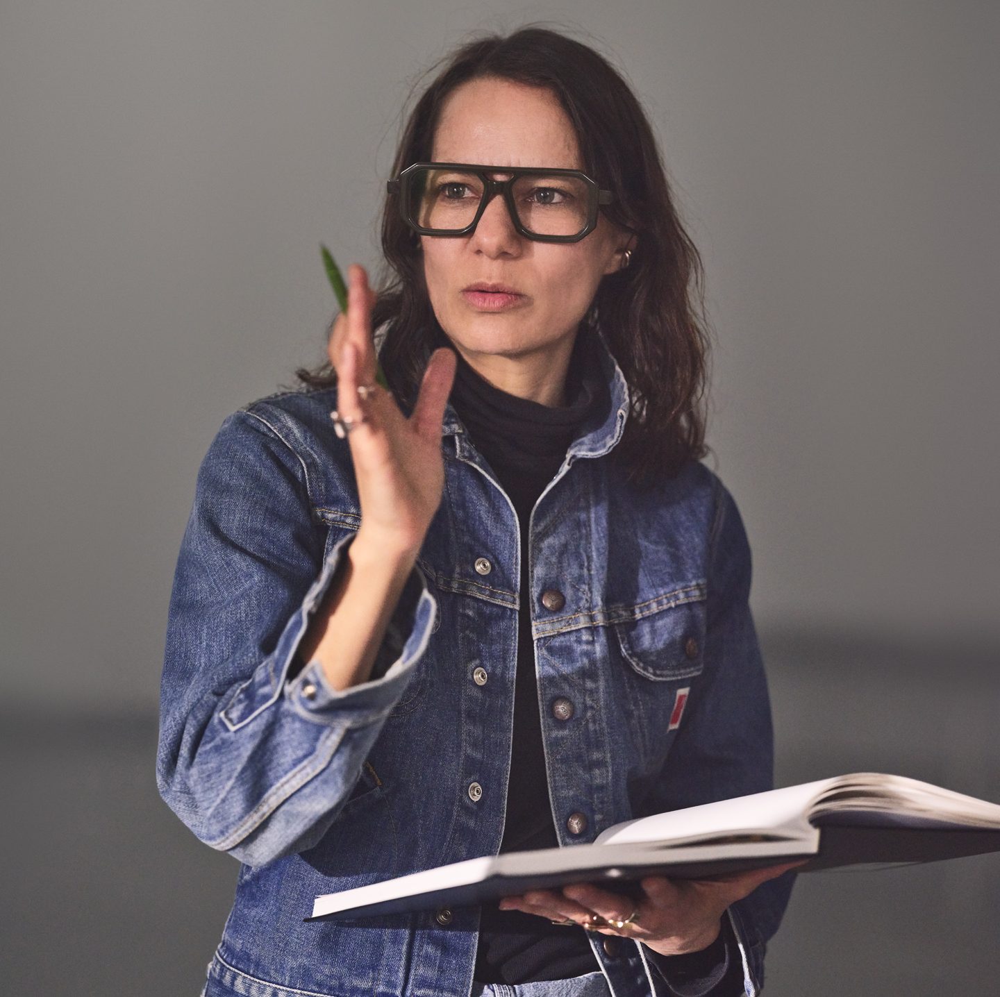 Headshot of Justine Chambers, looking just past the camera. She is wearing thick glasses and is holding a book open in one hand and a pen in other.