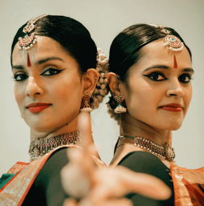 Two women in traditional bharatanatyam clothing, hair, makeup and jewelry pose for the camera. They are standing back to back, looking at the camera and are pointing their hands towards it.