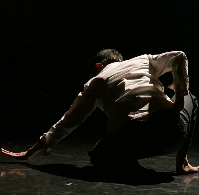 A dance artist crouches low to the stage with her back to the camera. One arm is extended out to the left.
