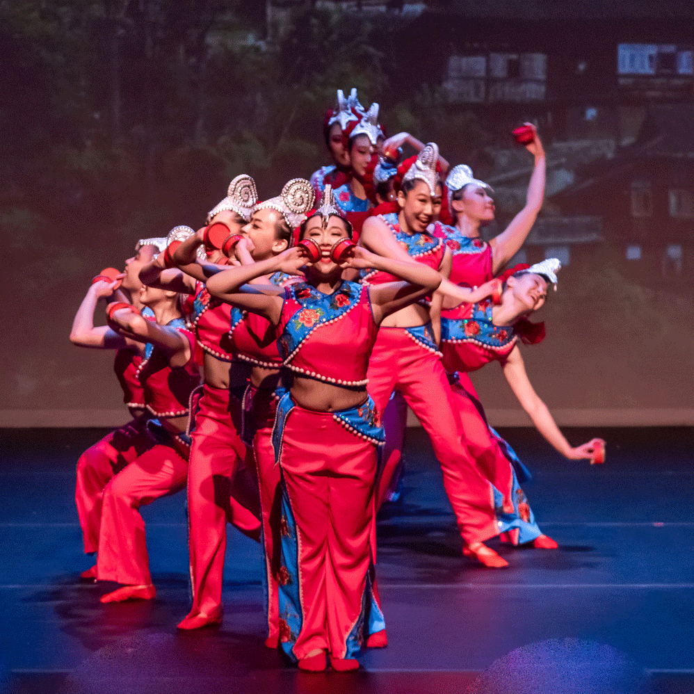 A group of young Asian dancers. They are wearing bright red outfits and are wearing silver helmets. They are standing in a line on the stage.