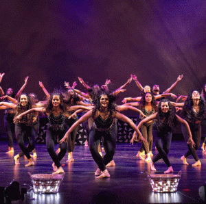 A large group of dancers on stage. They are wearing dark clothing and are standing in rows, bending over with their arms extended.